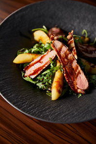 delicious warm salad with arugula, potato and meat in black plate on wooden table