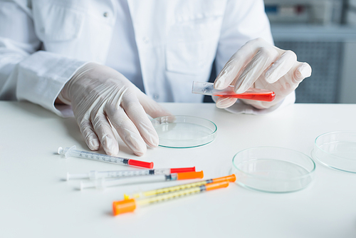 Cropped view of scientist in latex gloves holding test tube near petri dishes and syringes in lab