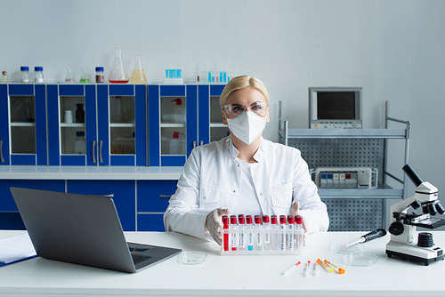 Scientist in protective mask looking at camera near test tubes and microscope in lab