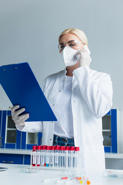 Scientist in protective mask talking on smartphone and holding clipboard near test tubes in lab