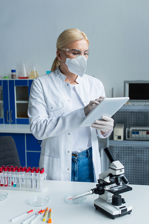 Scientist in protective mask using digital tablet near test tubes and microscope in lab