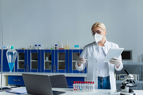 Scientist in protective mask holding petri dish and digital tablet in laboratory