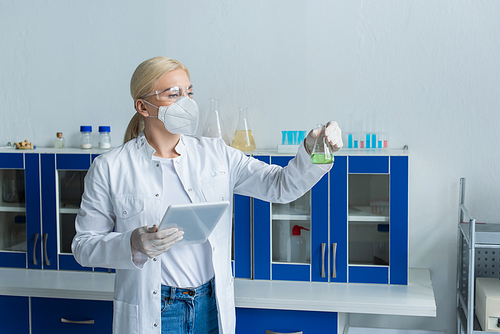 Researcher in protective mask holding flask and digital tablet in laboratory