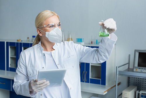 Scientist in protective mask holding flask with liquid and digital tablet in laboratory