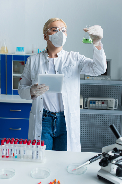 Scientist in protective mask holding flask and digital tablet near test tubes in lab