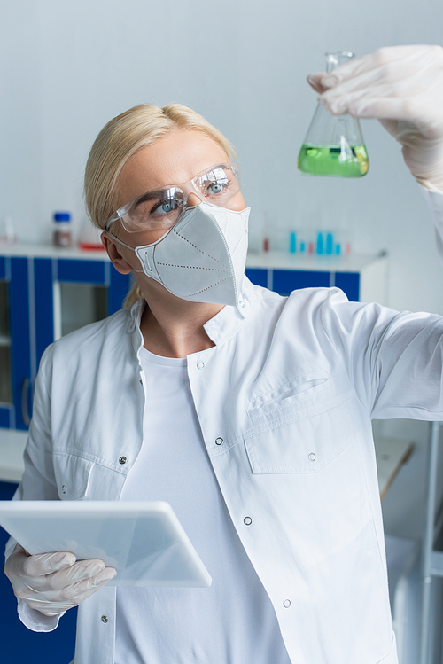 Scientist in goggles looking at blurred flask and holding digital tablet in lab