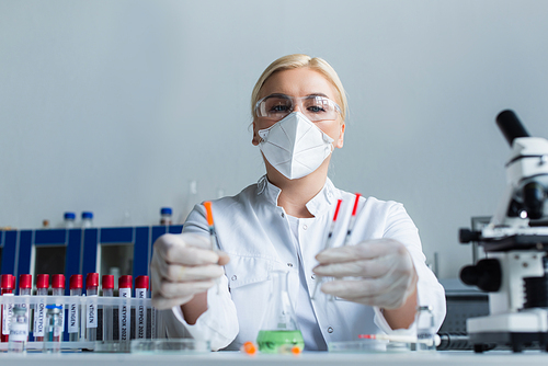 Low angle view of scientist in mask holding blurred syringes near microscope and test tubes in lab