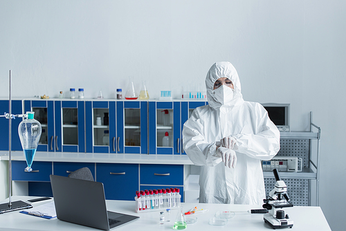 Scientist in hazmat suit and latex gloves standing near test tubes and microscope in lab