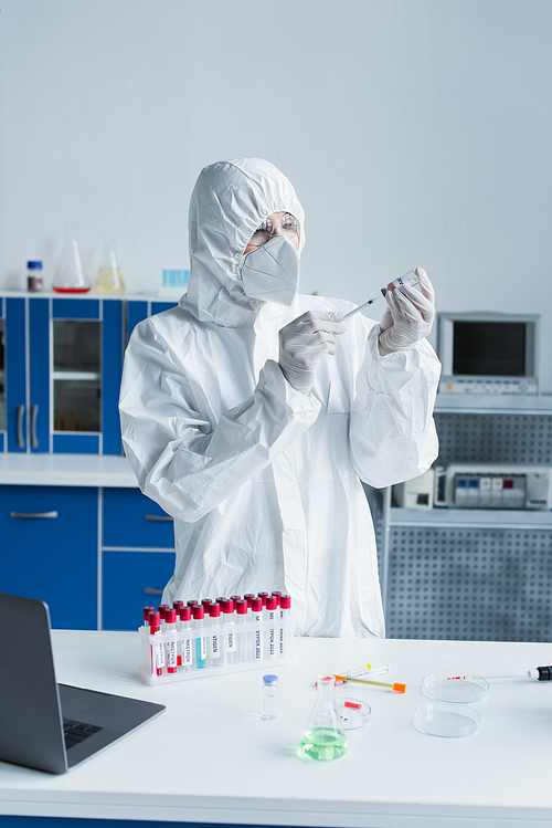 Scientist in hazmat suit holding syringe and vaccine near test tubes in lab