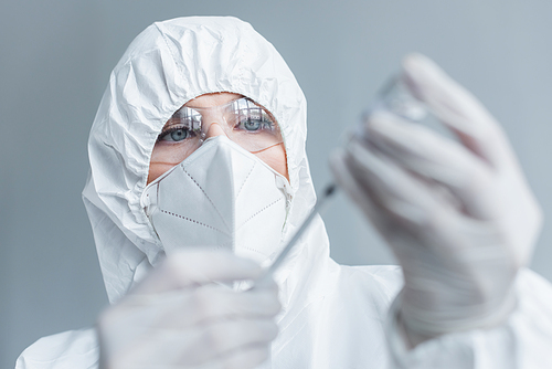 Scientist in hazmat suit and goggles holding blurred syringe and vaccine in lab