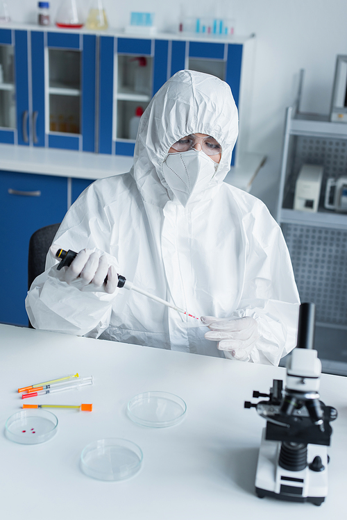 Scientist in latex gloves and hazmat suit holding electronic pipette and petri dish in lab
