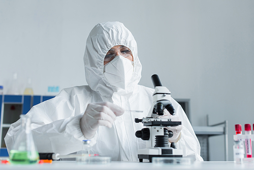 Scientist in hazmat suit holding glass while working with microscope in laboratory