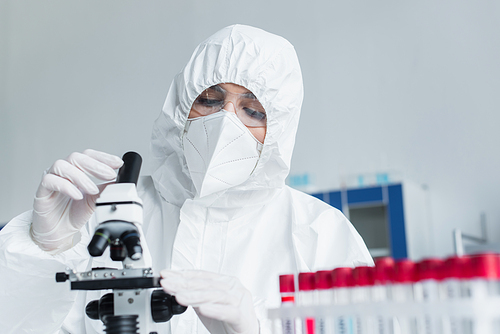 Scientist in hazmat suit using microscope near blurred test tubes in lab