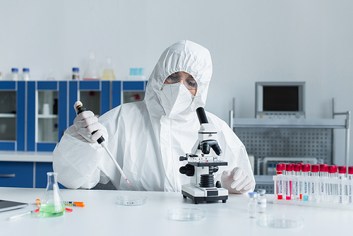 Scientist in protective suit holding electronic pipette near microscope and test tubes in lab