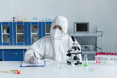 Scientist in hazmat suit writing on clipboard near microscope and test tubes in lab