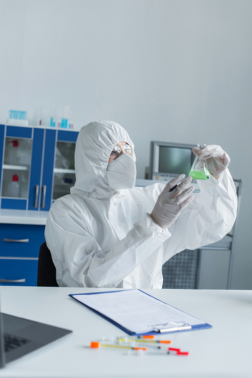 Scientist in hazmat suit holding flask near blurred clipboard and laptop in laboratory