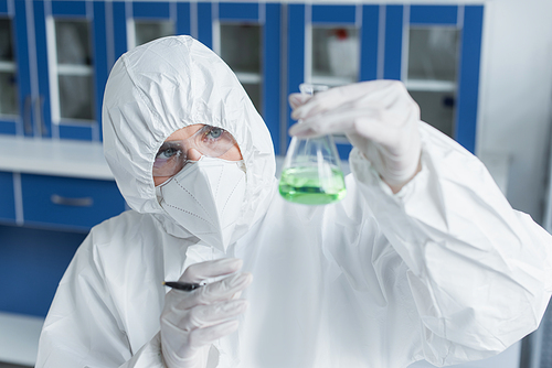 Scientist in protective suit and mask holding flask with liquid in lab