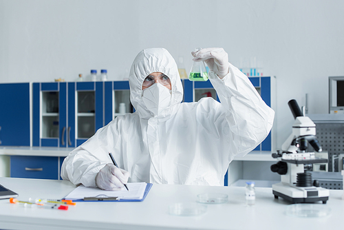 Scientist in hazmat suit working with flask and writing on clipboard in laboratory