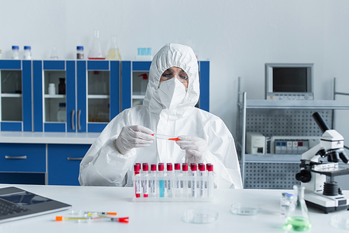 Scientist in protective goggles and suit holding test tube near laptop and microscope in lab