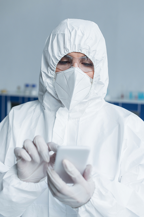 Scientist in hazmat suit and protective mask using blurred smartphone in lab