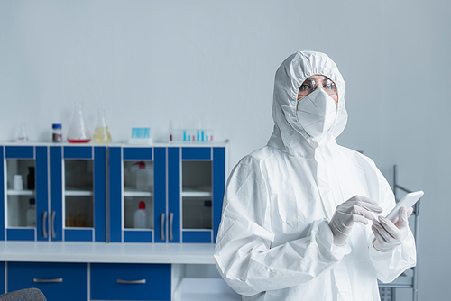 Scientist in protective suit and goggles holding smartphone and looking at camera in laboratory