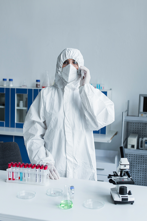Scientist in protective suit and mask talking on smartphone near test tubes and microscope in lab