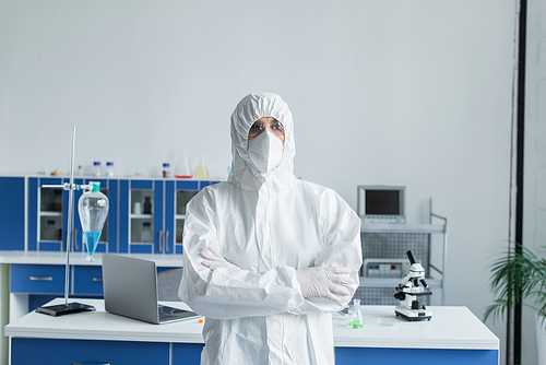 Scientist in hazmat suit and protective goggles crossing arms in lab