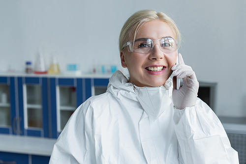 Smiling scientist in protective suit talking on smartphone in lab