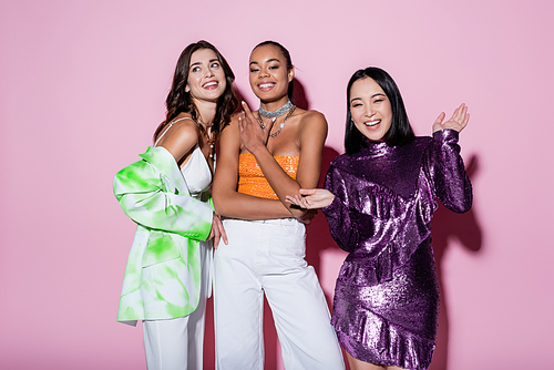 happy multiethnic women in trendy outfits posing on pink