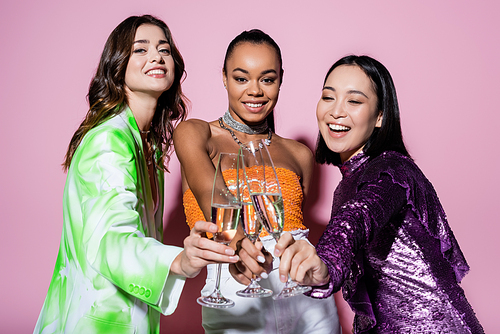 cheerful interracial women in trendy outfits clinking glasses of champagne on pink