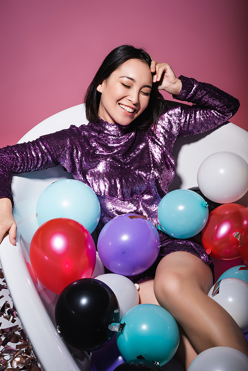 happy asian woman in purple dress lying in bathtub with colorful balloons on pink