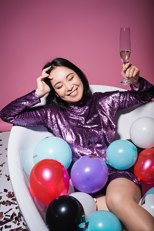 happy asian woman in purple dress lying in bathtub with colorful balloons and holding glass of champagne on pink