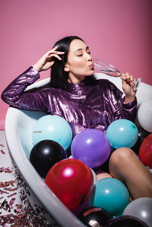 asian woman in purple dress lying in bathtub with colorful balloons and drinking champagne on pink