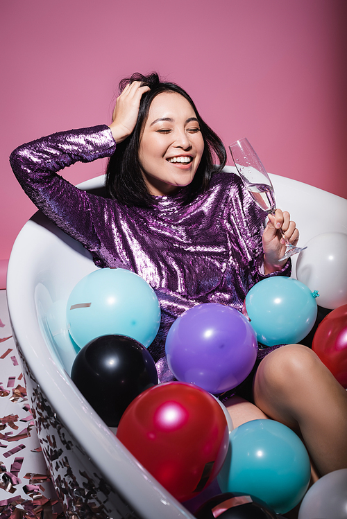 cheerful asian woman in purple dress lying in bathtub with colorful balloons and holding glass of champagne on pink