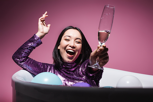 excited asian woman in purple dress lying in bathtub with balloons and holding glass of champagne on pink