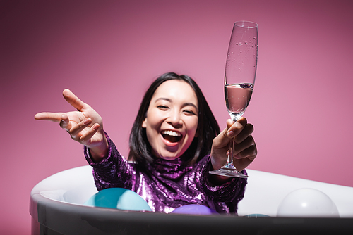 amazed asian woman in purple dress lying in bathtub with balloons and holding glass of champagne on pink