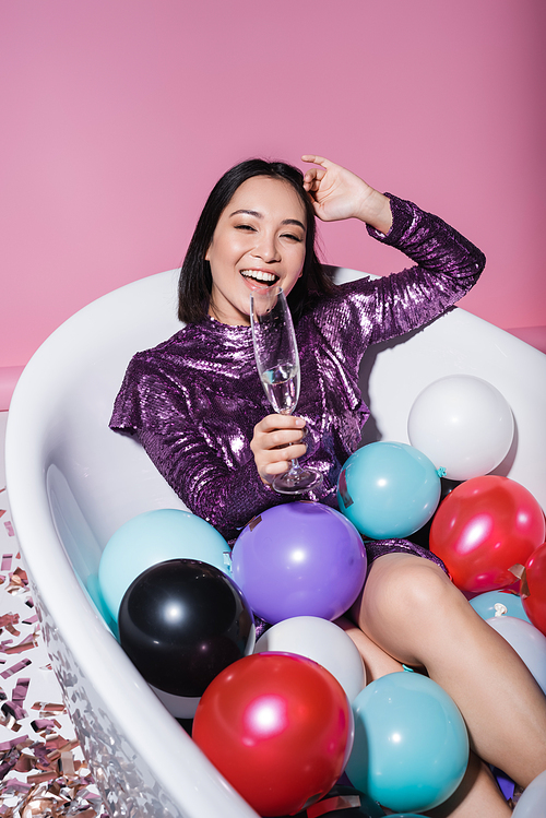 amazed asian woman in dress lying in bathtub with colorful balloons and holding glass of champagne on pink
