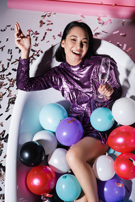 high angle view of happy asian woman lying in bathtub with colorful balloons and holding glass of champagne