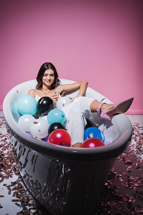 happy woman lying in bathtub with colorful balloons and holding glass of champagne on pink