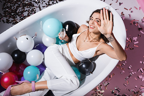 top view of happy woman with tattoo gesturing and lying in bathtub with balloons while holding glass of champagne