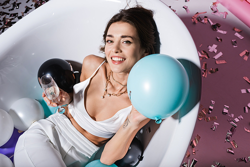 top view of cheerful woman with tattoo gesturing and lying in bathtub with balloons while holding glass of champagne