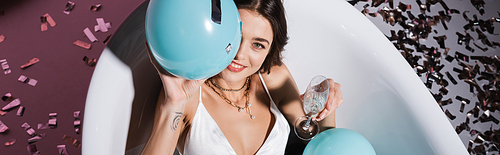 top view of smiling woman with tattoo lying in bathtub while holding balloon and glass of champagne, banner
