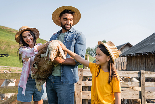 woman and child in straw hats stroking lamb in hands of smiling farmer