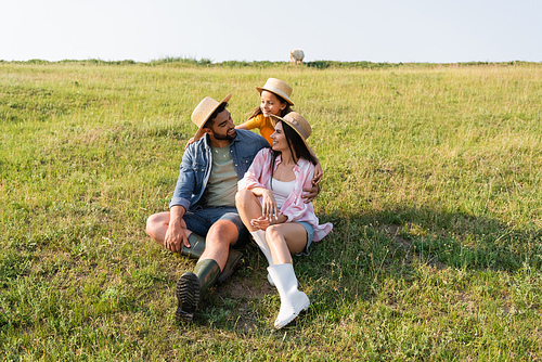 joyful family in straw hats embracing in grassy meadow on farmland