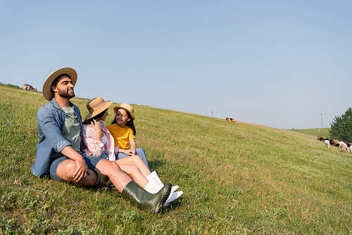 happy farmer looking away while sitting in field near family under blue sky