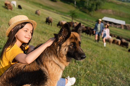 happy girl in straw hat petting cattle dog near blurred parents in green meadow