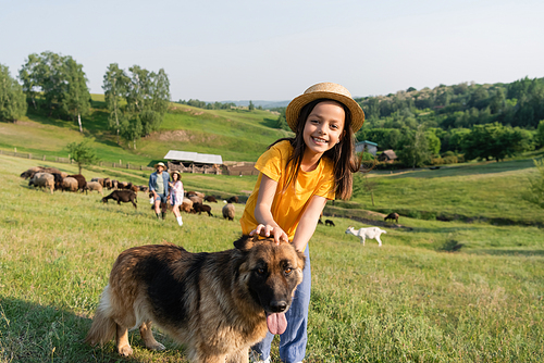 joyful girl looking at camera near cattle dog and parents herding flock on blurred background