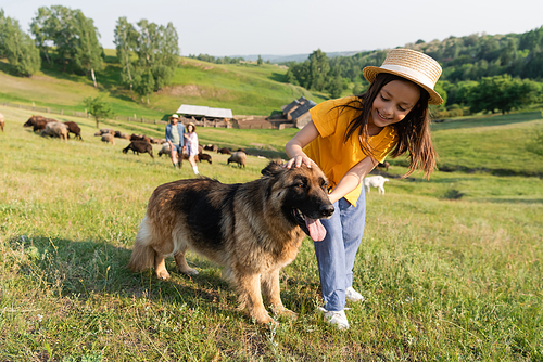 girl in straw hat cuddling cattle dog near parents herding livestock on blurred background