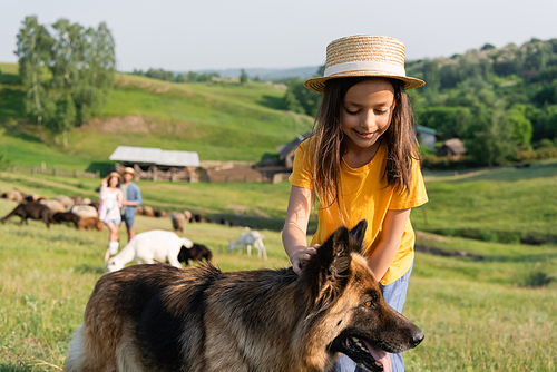 smiling child in straw hat stroking cattle dog near parents herding flock on blurred background