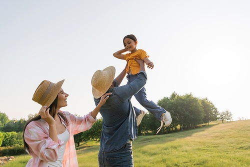 man in straw hat raising up cheerful daughter near happy wife